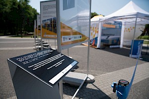 plansza dla osób niewidomych z informacją o wystawach, w tle wystawa i namioty wystawiennicze oraz wstęga w tle