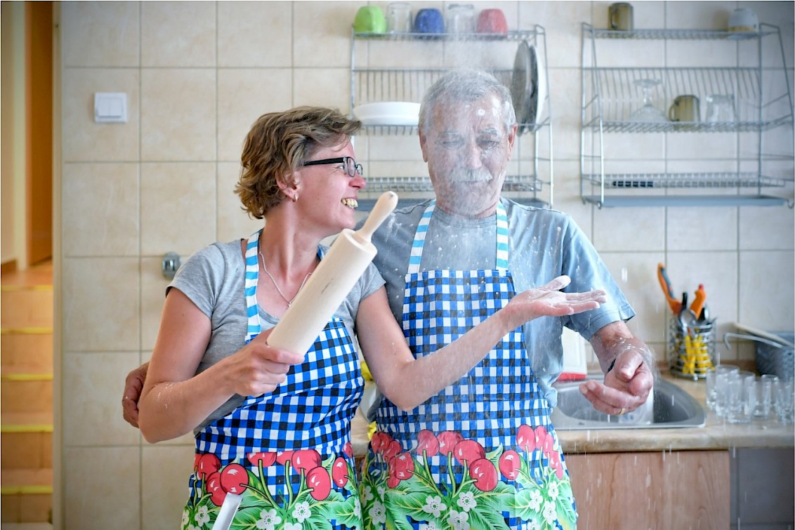 Obraz przedstawia starszą parę w kuchni. Kobieta rozsypuje mąkę na mężczyznę.