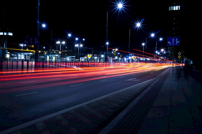 Obraz przedstawia ulicę nocą. Wyróżnione na obrazie są światła po autach oraz oświetlenie uliczne