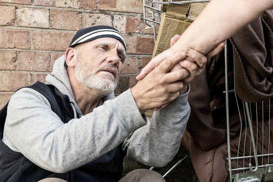 Obraz przedstawia osobę bezdomną trzymającą za rękę inną osobę