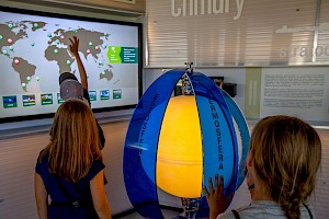 trójka dzieci odwrócona tyłem korzystająca z interaktywnego monitora, na którym wyświetlona jest mapa świata