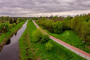Niewielka rzeka obok, której (z prawejstrony)  biegnie ścieżka rowerowa. Po obu stronach zielona roślinność.