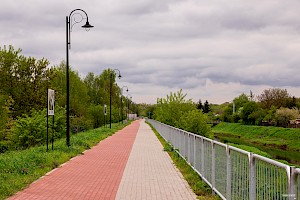 Widok na ścieżkę rowerową ograniczoną metalową barierką z prawej stronu poza barierką płynie rzeka. Po lewej stronie rząd latarni