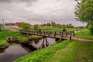 Drewniany most na niewielkiej rzece. Na moście z lewej strony porusza się rowerzystka. Naprzeciw niej pieszo idzie mężczyzna prowadzący psa na smyczy.