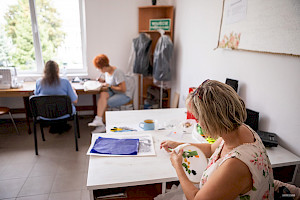 Trzy kobiety podczas zajęć integracyjnych. Na pierwszym planie osoba zwrócona bokiem zajmuje się wyszywaniem.